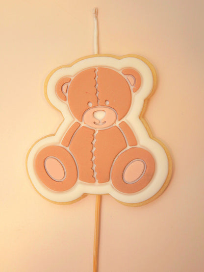 Teddy bear-shaped cookie on a stick with a wick against a beige background