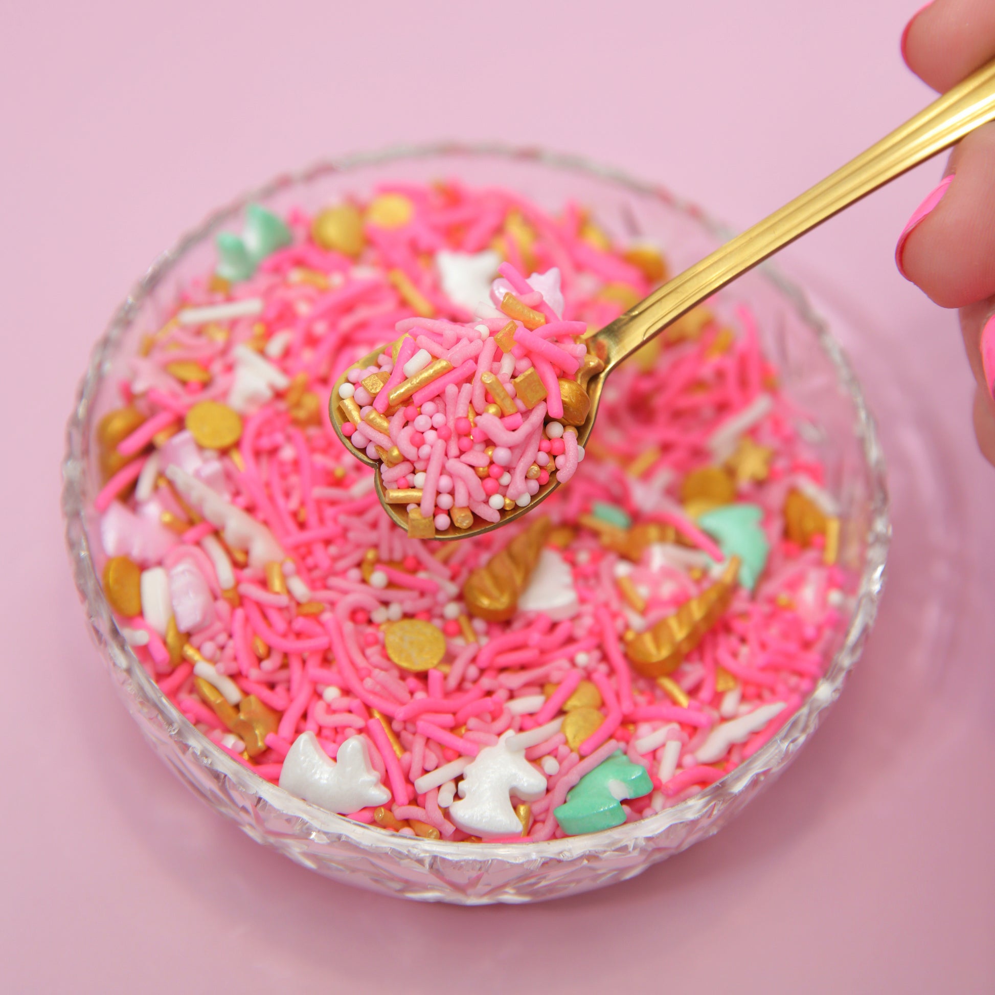 Glass bowl filled with bright pink sprinkles and a gold spoon on a pink background