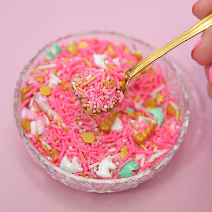 Glass bowl filled with bright pink sprinkles and a gold spoon on a pink background