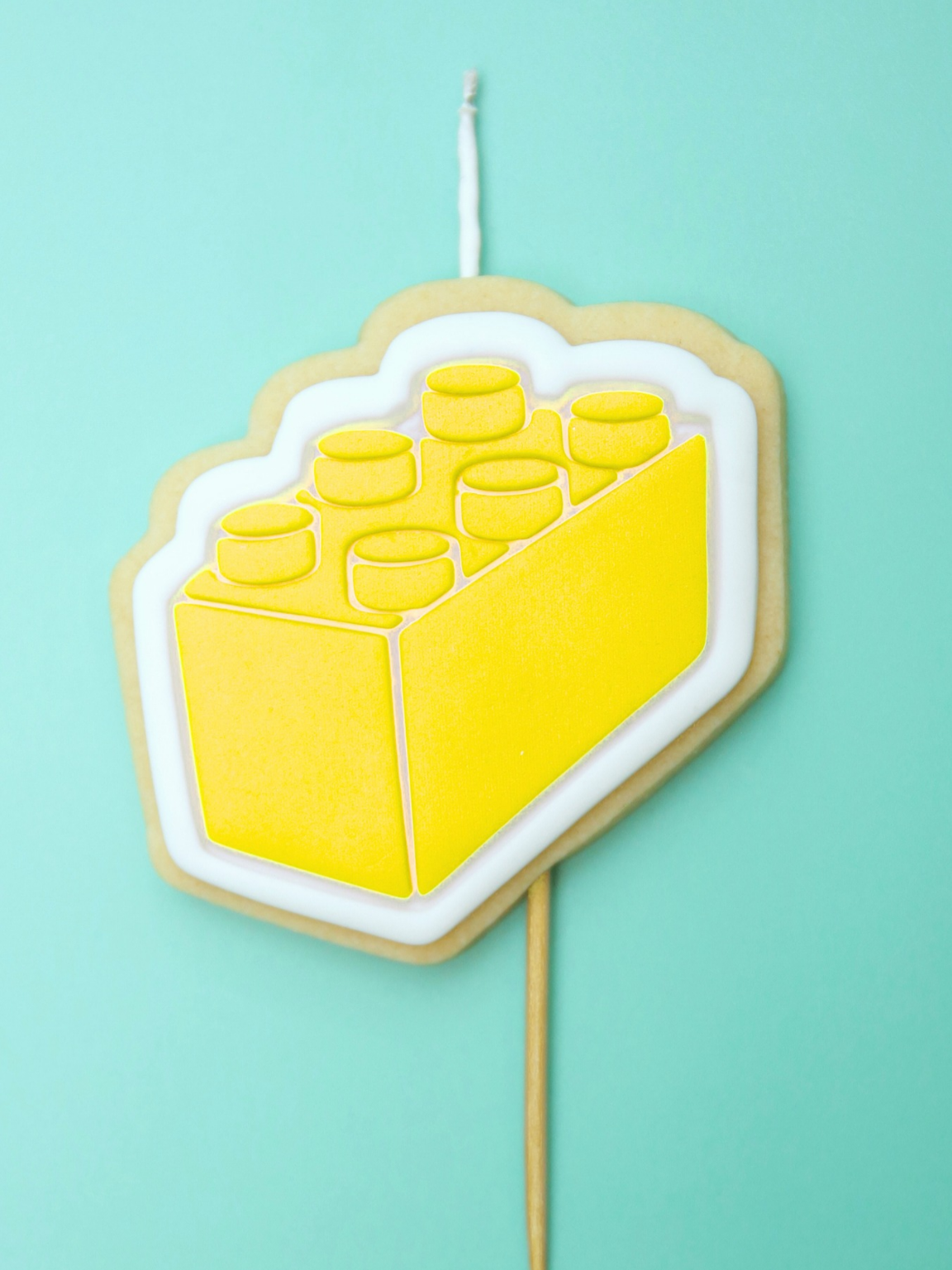 Yellow block-shaped cookie on a stick with a wick against a light blue background