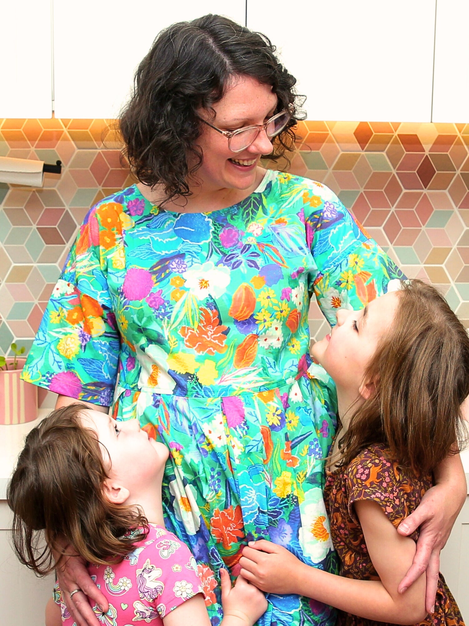 Woman in a colorful dress holding two children in a kitchen with a patterned backsplash.
