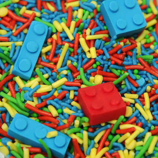 A close-up of colorful sprinkles mixed with edible toy building block pieces, including blue and red bricks, in bright red, blue, yellow, and green.