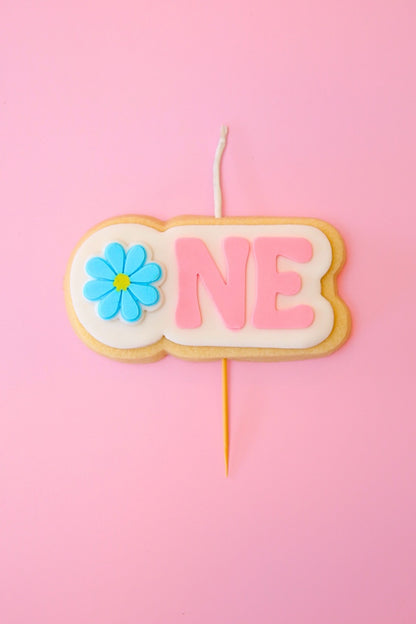 Candle shaped like a cookie with a flower and 'ONE' on a pink background