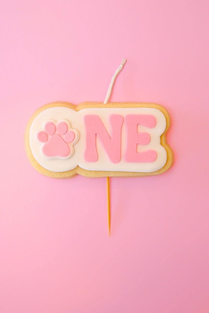 Candle shaped like a cookie with 'ONE' and a paw print on a pink background