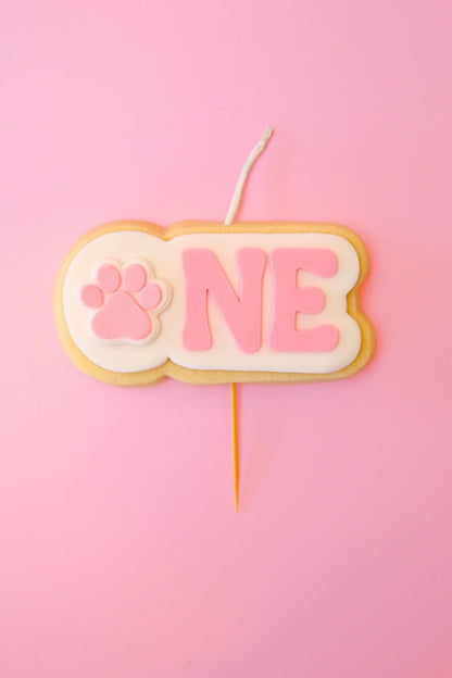 Candle shaped like a cookie with 'ONE' and a paw print on a pink background