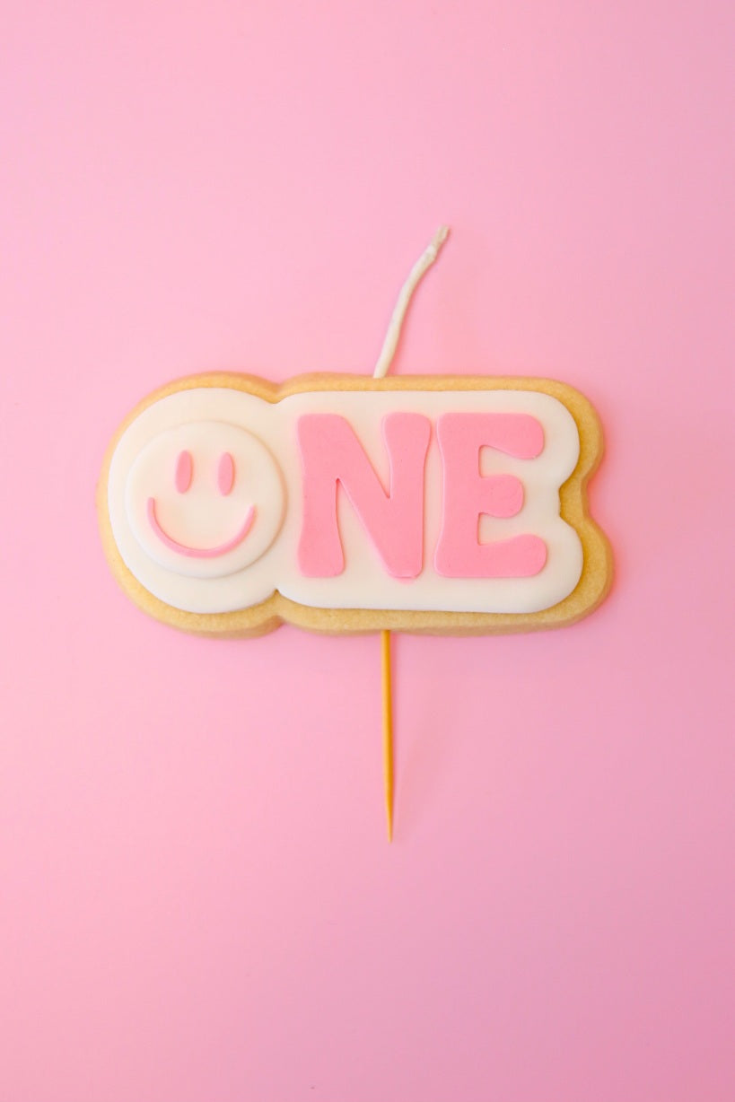 Candle shaped like a cookie with 'ONE' and a smiley face on a pink background