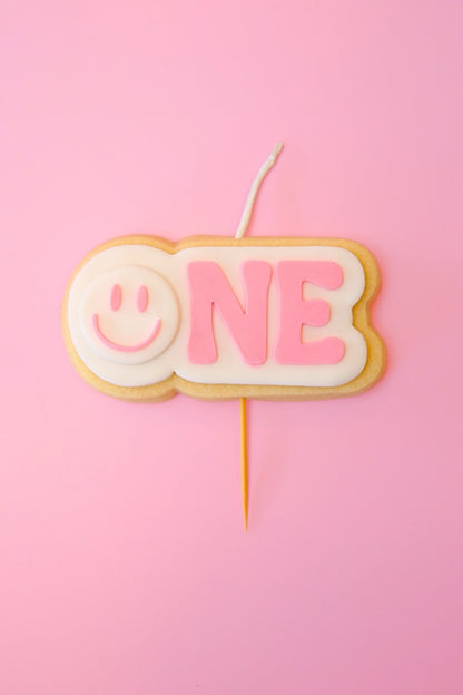 Candle shaped like a cookie with 'ONE' and a smiley face on a pink background
