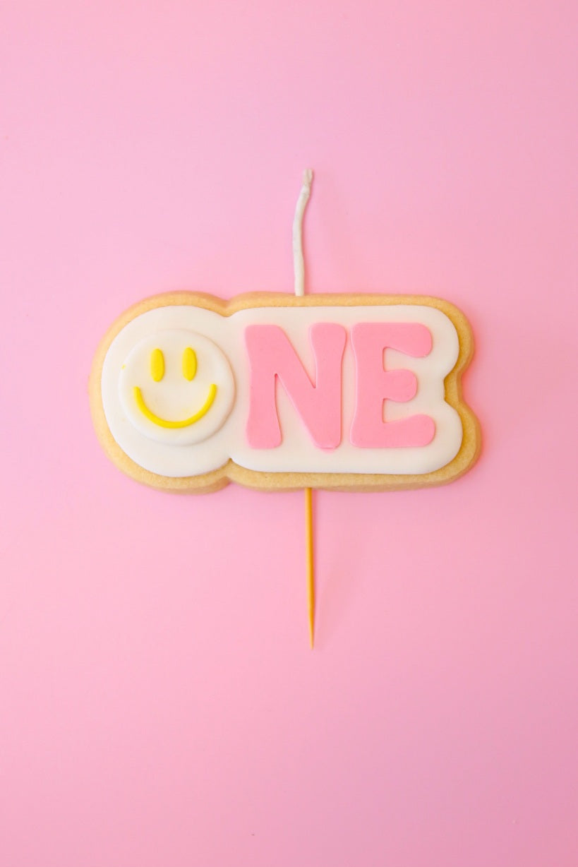 Candle shaped like the number '1' with a yellow smiley face on a pink background
