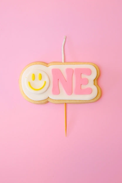 Candle shaped like the number '1' with a yellow smiley face on a pink background