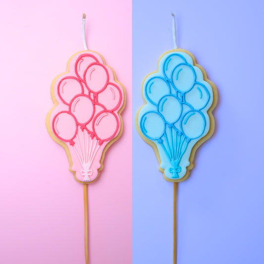 Two balloon-shaped cookie candles, one pink and one blue, on a two-tone pink and purple background.