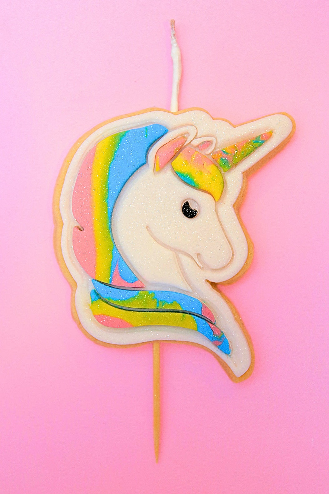 Unicorn shaped cookie with a wick on a stick with rainbow hair against a purple background