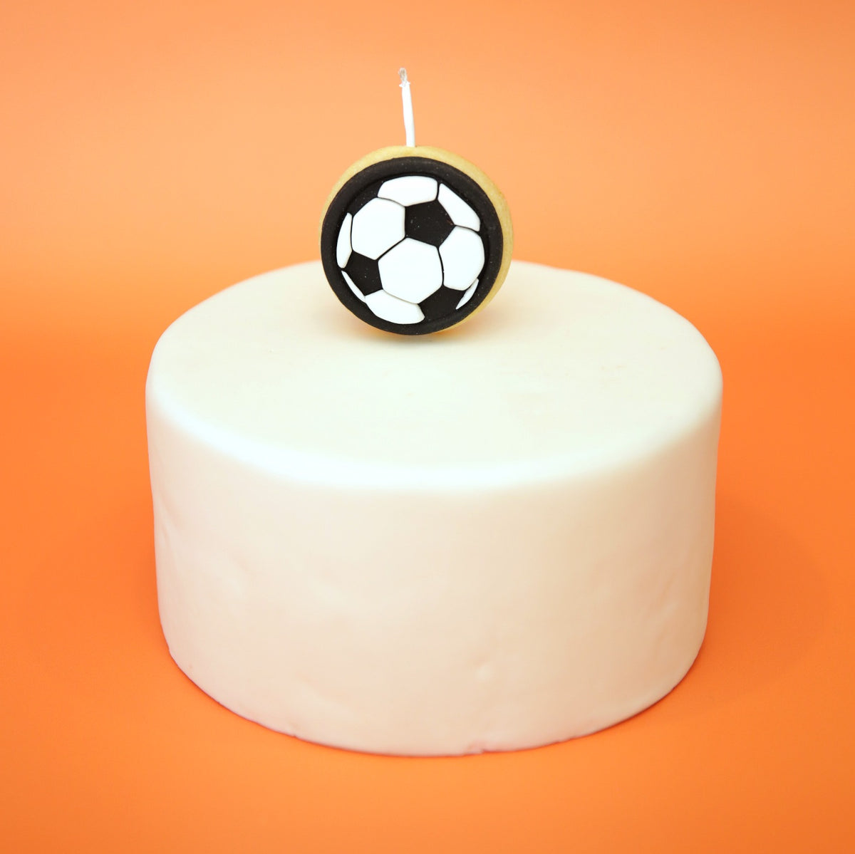A white frosted round cake topped with a black and white soccer ball cookie candle on a solid orange background.