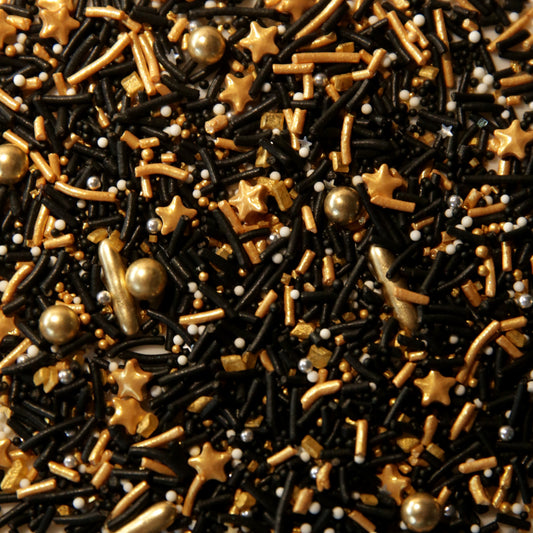 A close-up of mixed sprinkles and candy decorations in black, metallic gold, and small white pearls, filling the entire frame.
