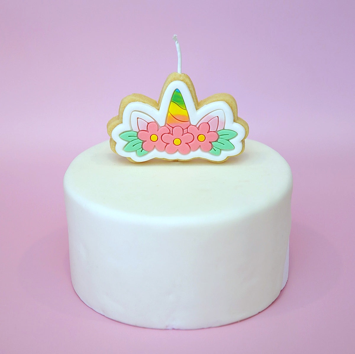 A unicorn crown shaped cake topper candle with pink flowers, green leaves, and a rainbow horn, displayed on top of a round white frosted cake against a soft pink background.